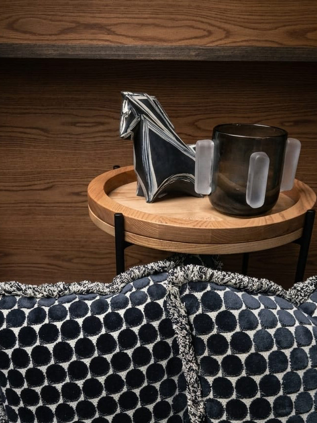 Two dark throw pillows with a black and beige geometric pattern and fringed edges, styled on a chair in front of a wooden side table holding a horse figurine and a dark grey glass vase.